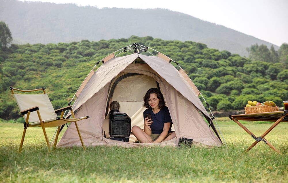Femeie cu telefon în cort lângă un aparat de aer condiționat la un camping cu scaun și masă