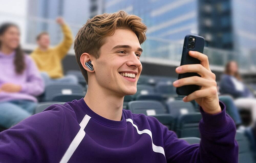 Un tânăr într-un hanorac violet stă pe tribune, zâmbind și ținând un smartphone cu o cască wireless în ureche