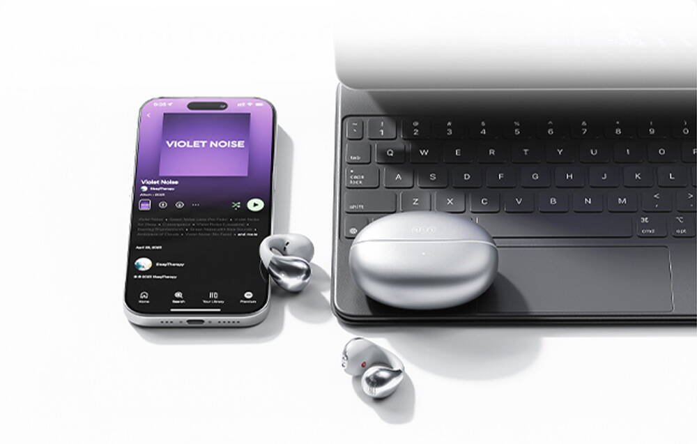 Căști wireless argintii și carcasă lângă un smartphone cu aplicația de muzică deschisă și o tastatură de laptop