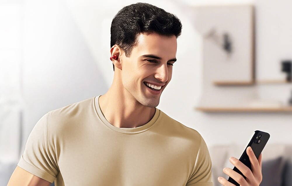 Un bărbat în cămașă bej zâmbește în timp ce se uită la telefonul său cu o cască roșie în ureche